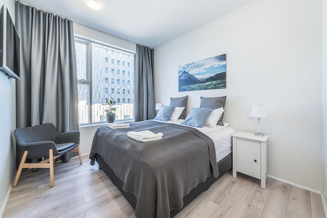 A modern bedroom with a large bed, grey curtains, a grey armchair, a small bedside table, and a window with a cityscape view.