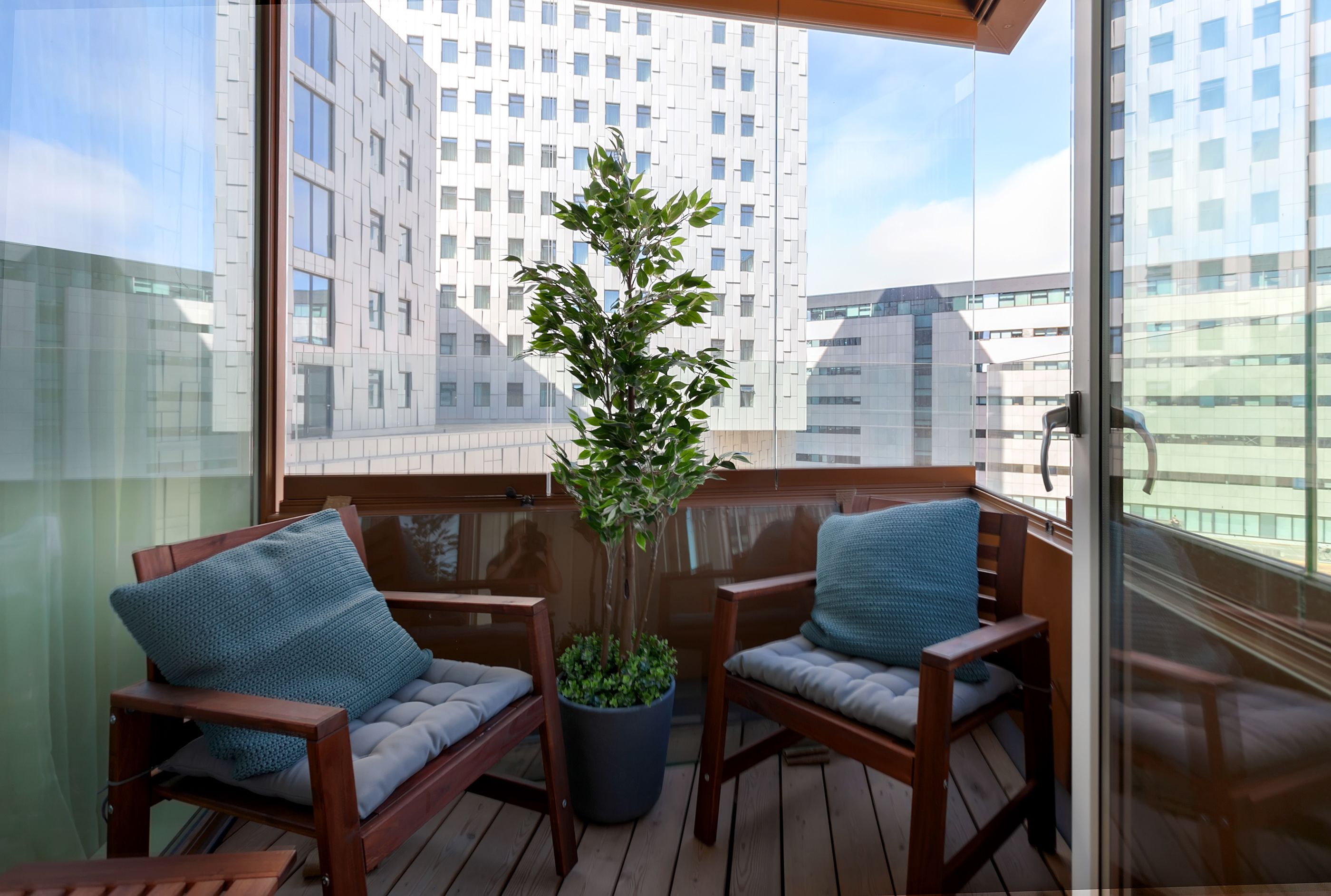 Un balcón con dos sillas de madera, una planta en maceta y vistas a edificios modernos.