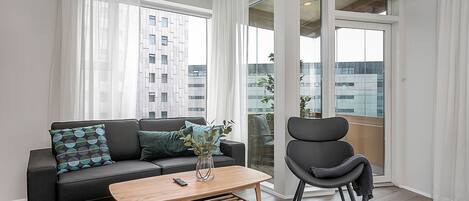 A modern living room with a black sofa, a wooden coffee table, and a black armchair. There is a large window with white curtains and a view of buildings outside.
