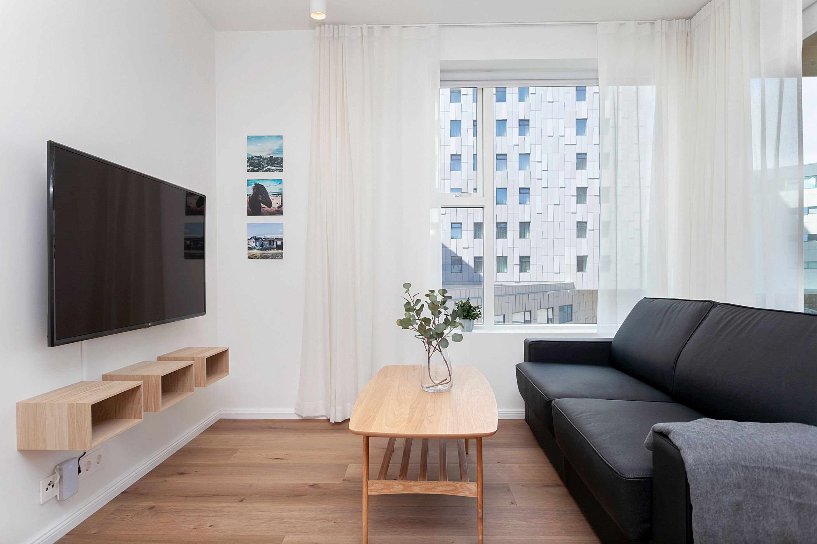 A modern living room with a flat-screen TV, wooden shelves, a coffee table with a vase, a black sofa, and a window with a city view.