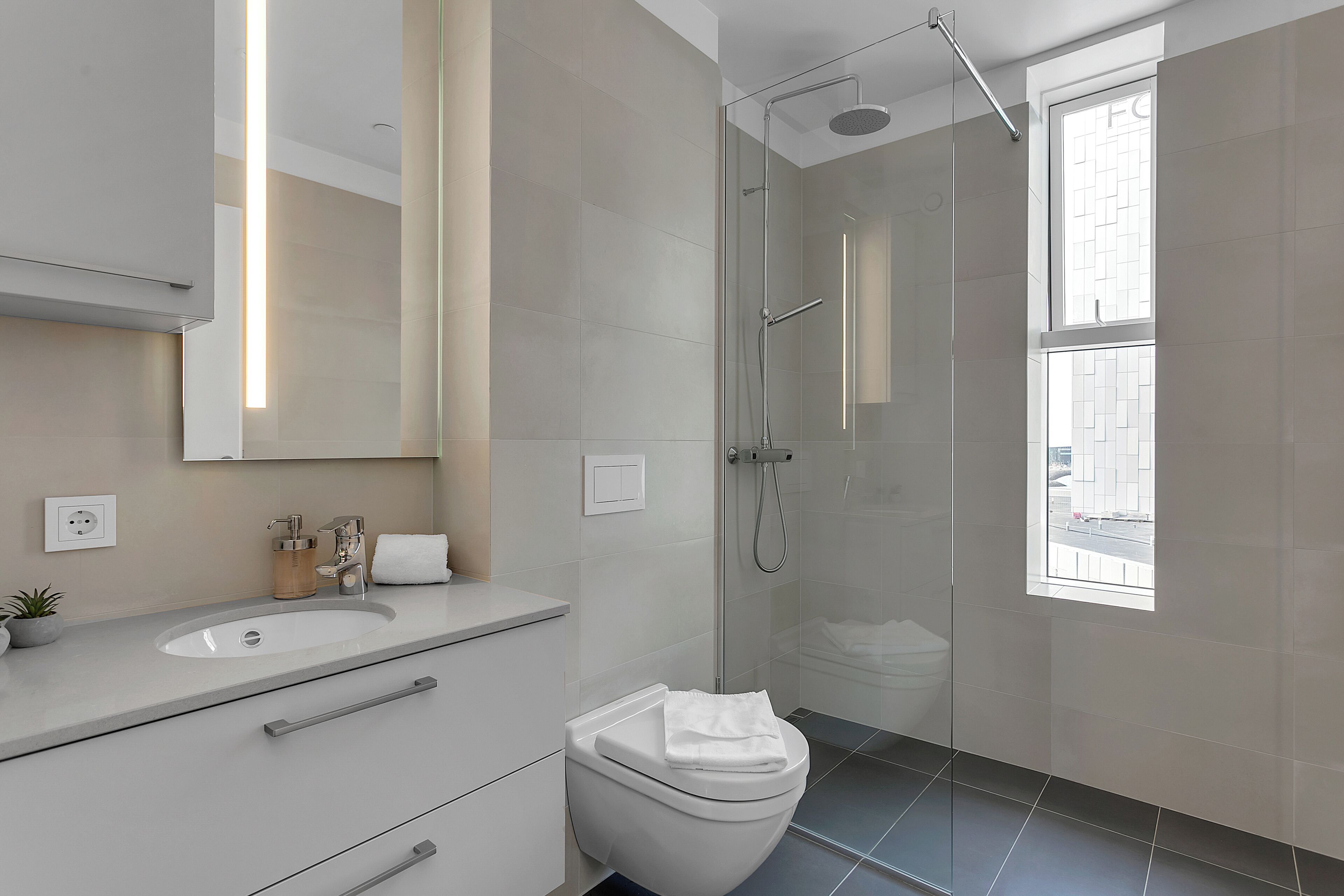 A modern bathroom with a glass-enclosed shower, a large mirror, and a window providing natural light.
