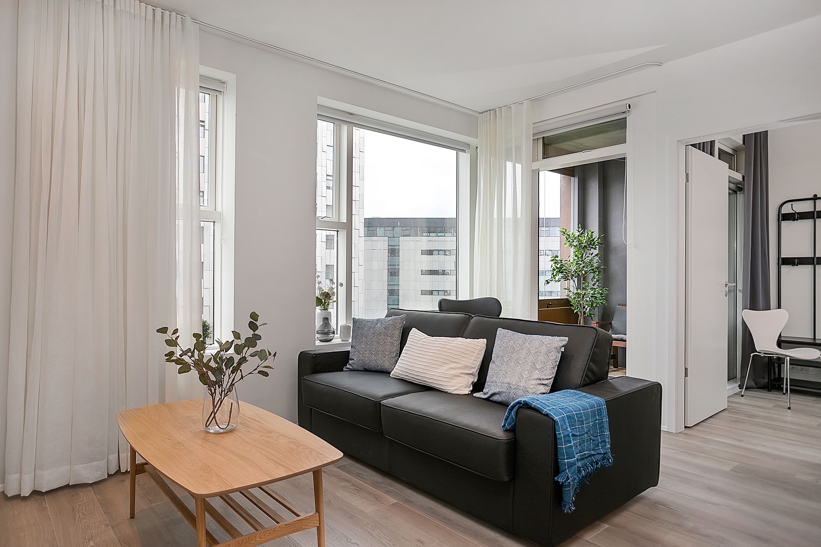 A modern living room with a black sofa, a wooden table, and a view of the city through large windows.