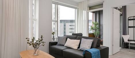 A modern living room with a black sofa, a wooden table, and a view of the city through large windows.