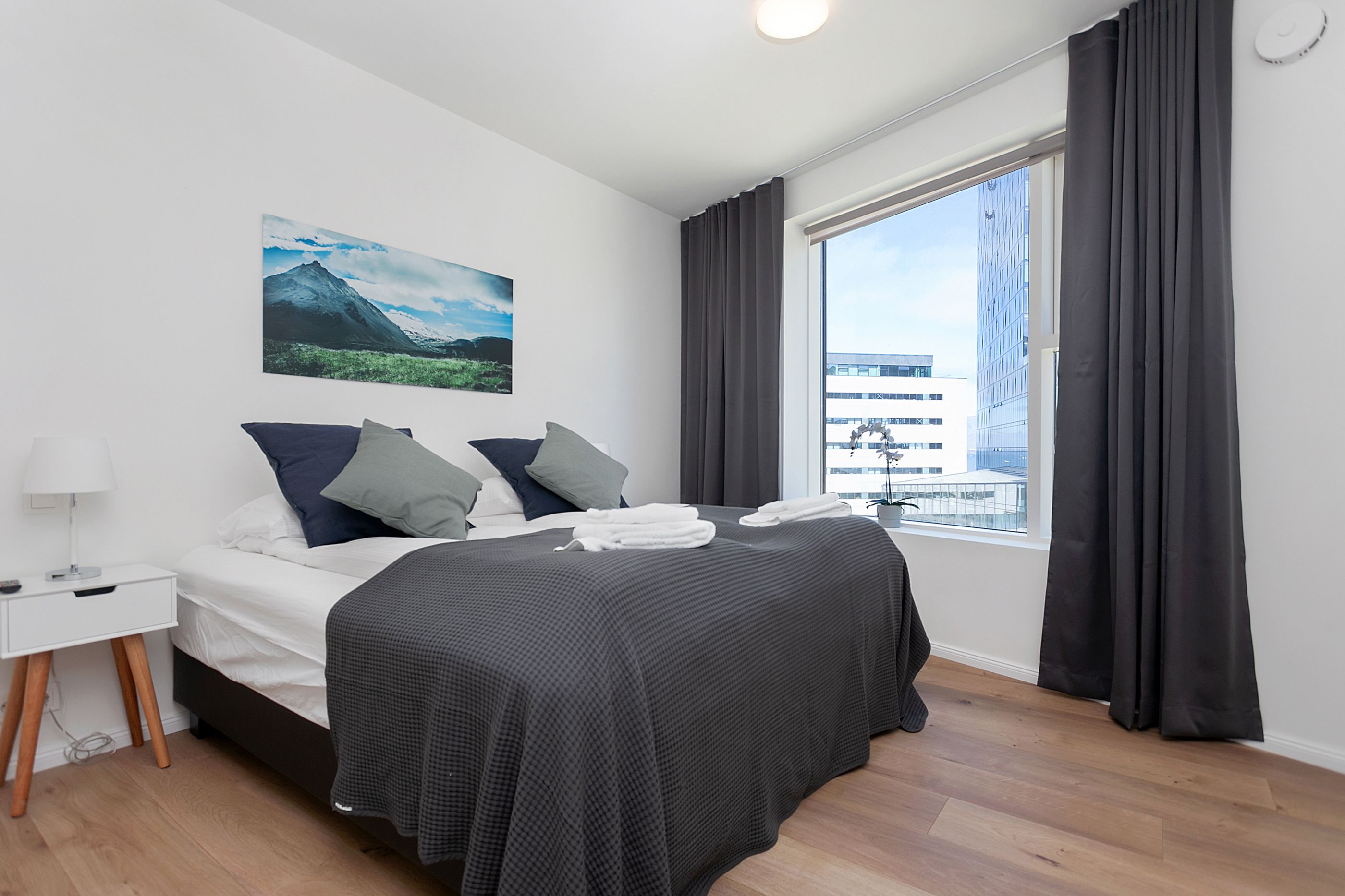 A modern hotel room with a large bed, a nightstand, a window with curtains, and a mountain landscape painting on the wall.