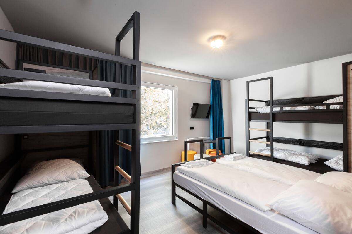 A dorm-style room with bunk beds, a window with curtains, a flat-screen TV, and a small orange chair.