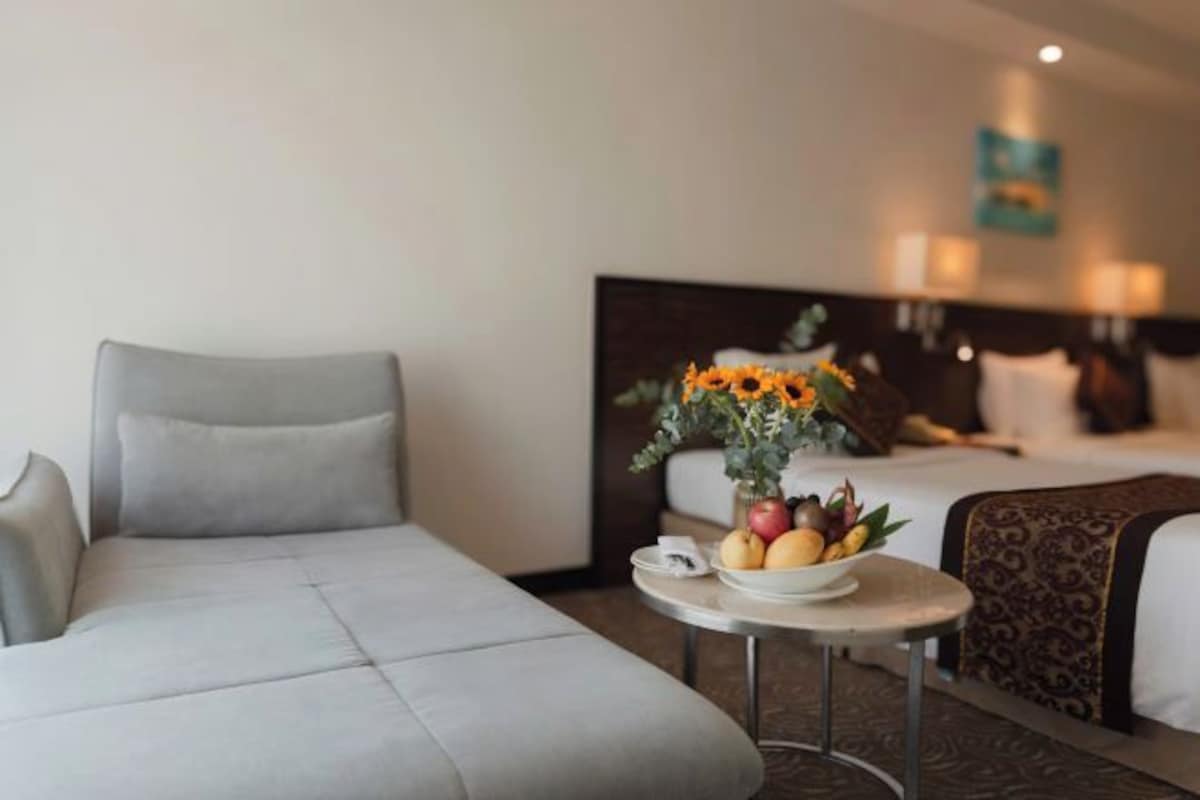 Une chambre d’hôtel avec un canapé gris, une petite table ronde sur laquelle se trouve un panier de fruits, et un lit doté d’une tête de lit.