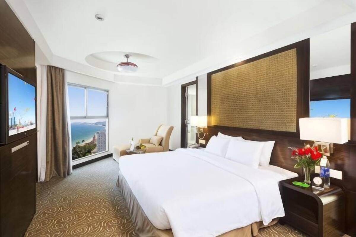 Ein Hotelzimmer mit einem großen Bett, einem Fernseher, einem Schreibtisch mit Lampe und Blick auf die Stadt.