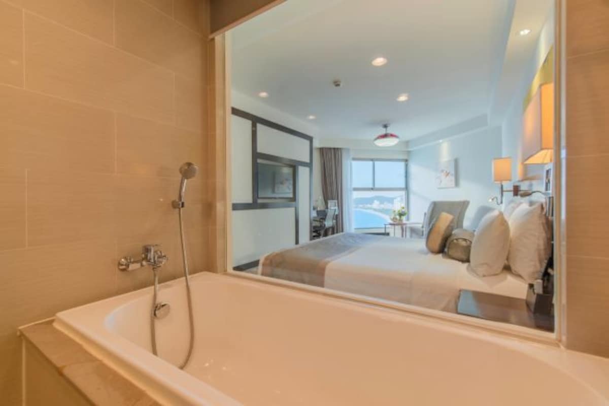 Une salle de bain moderne avec une grande baignoire et un miroir qui reflète une chambre à coucher avec un lit et une fenêtre.