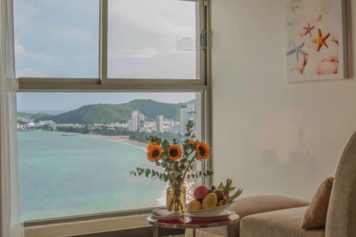 Une pièce dotée d’une grande fenêtre donnant sur une plage et sur le paysage urbain, un vase rempli de tournesols et un panier de fruits posé sur une petite table.
