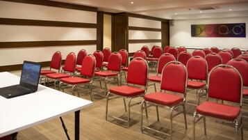 Un salón de conferencias con filas de sillas rojas, una mesa blanca con una computadora portátil y suelo de madera.