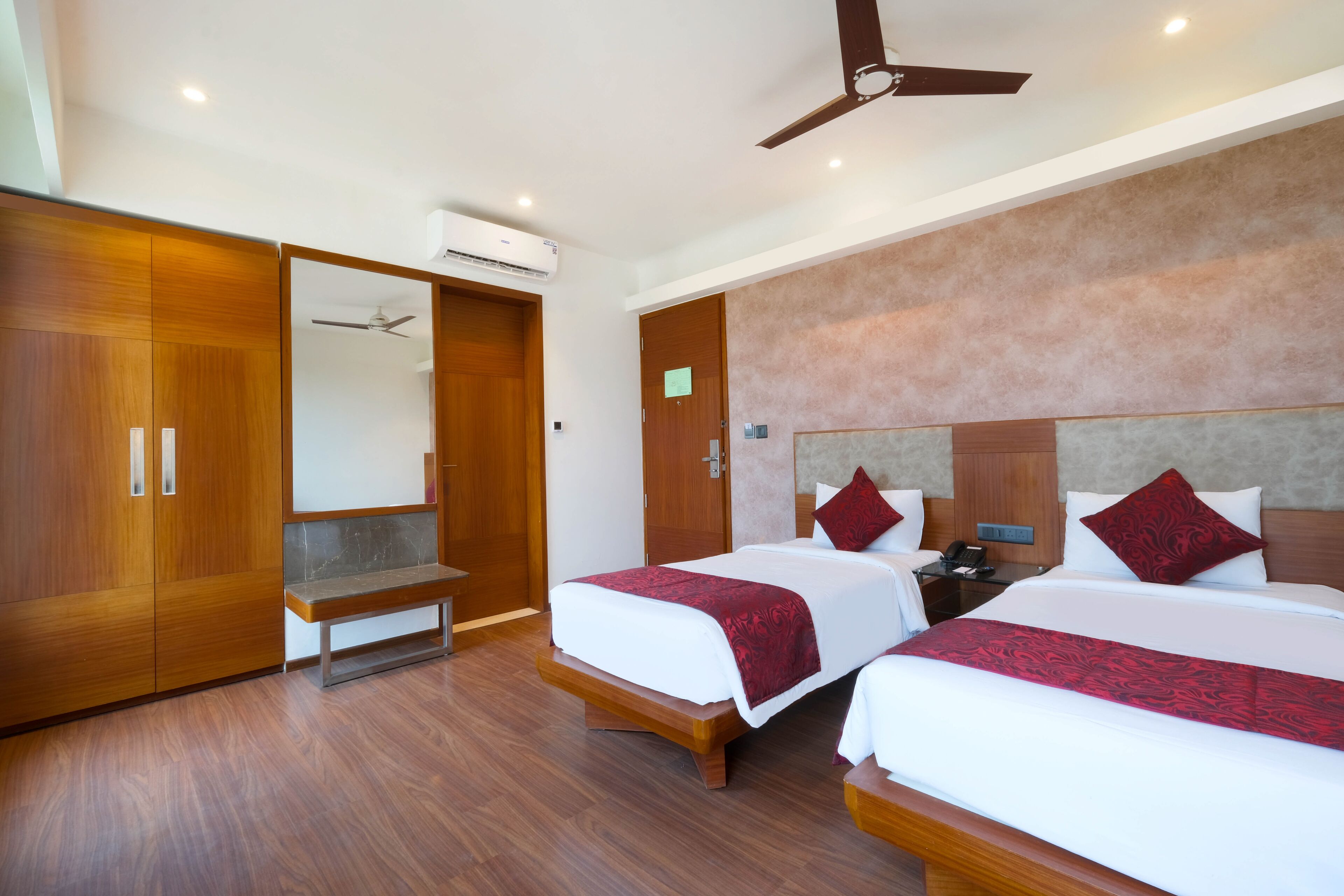 A hotel room with two beds, wooden flooring, a ceiling fan, and an air conditioning unit.
