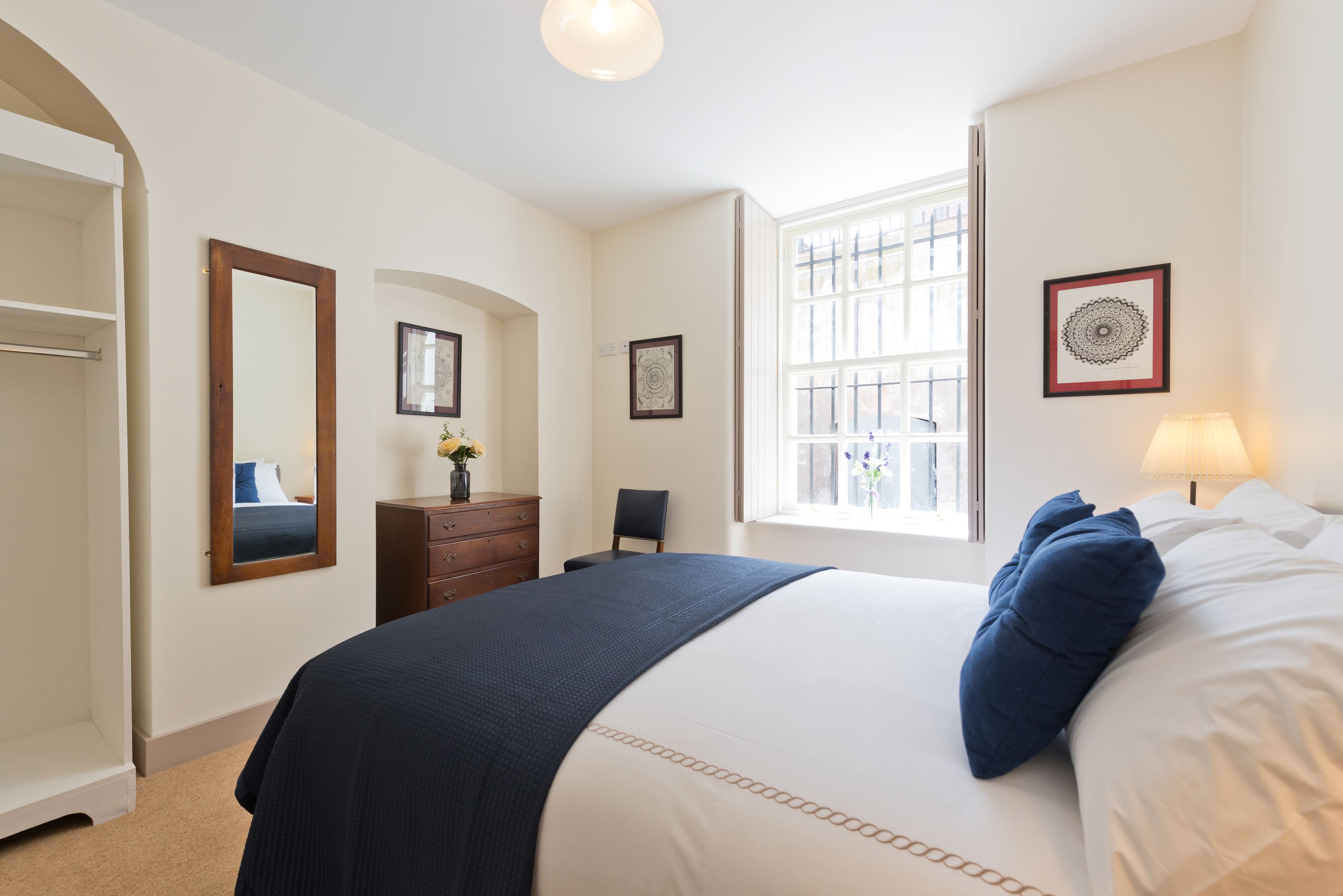 Een slaapkamer met een groot bed, een dressoir, een stoel, een raam met jaloezieën en een spiegel.