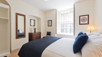 Een slaapkamer met een groot bed, een dressoir, een stoel, een raam met jaloezieën en een spiegel.