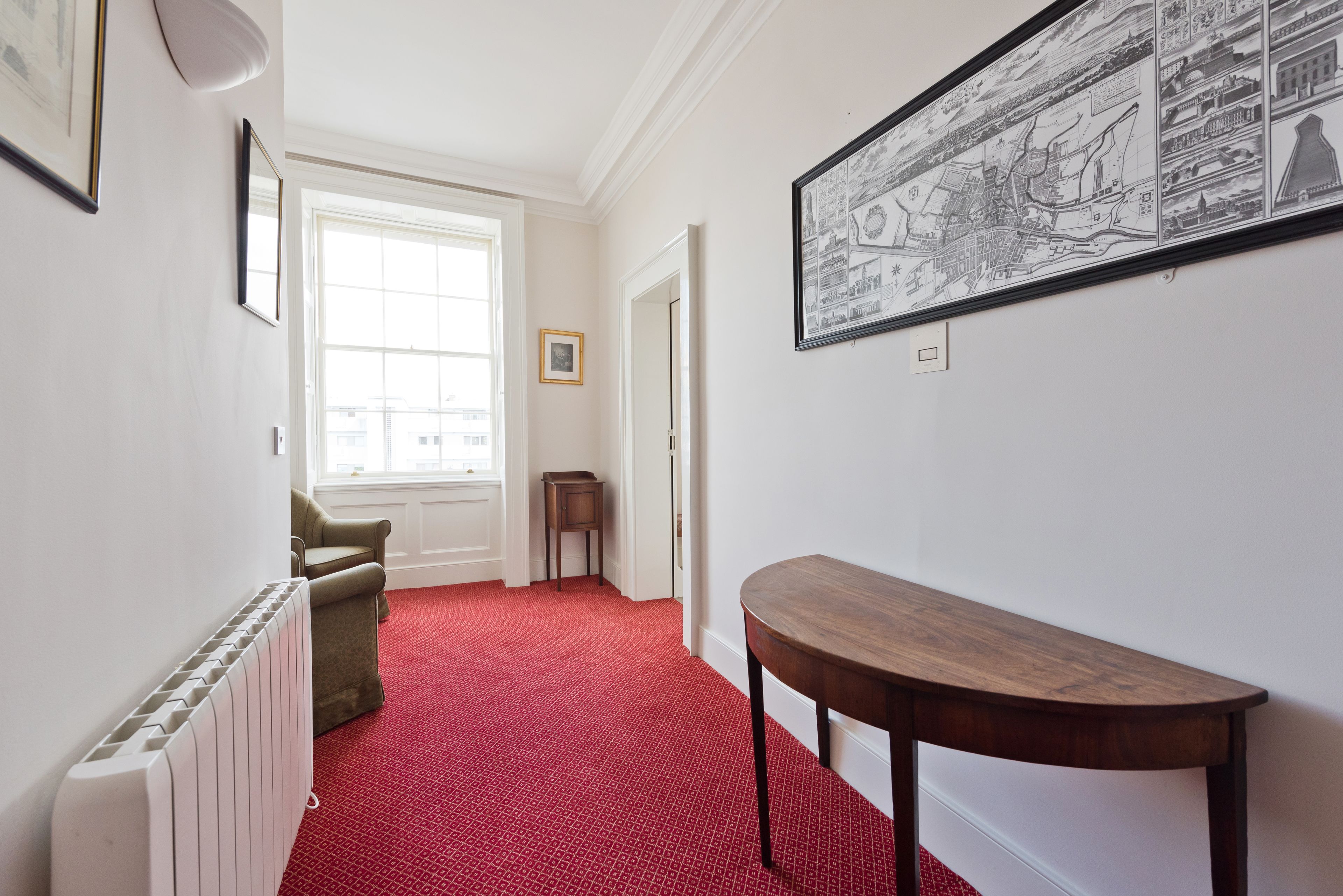 A hallway with a red carpet, a radiator, a green armchair, a wooden table, and a large black-and-white map on the wall.