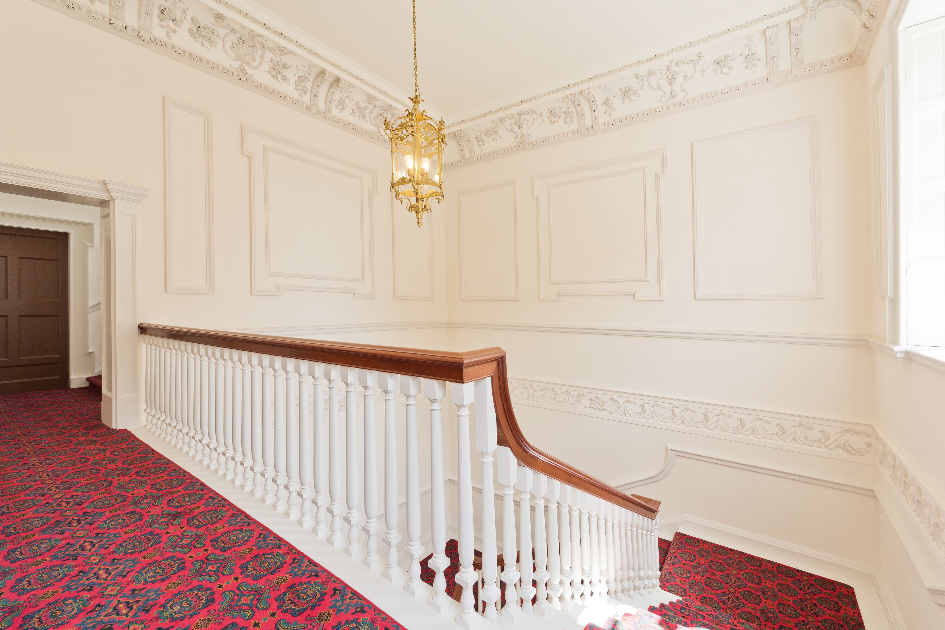Un magnifique escalier avec des balustrades blanches et une moquette rouge, menant à une pièce aux murs blancs richement décorés et ornée d’un lustre.