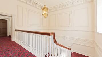 Un magnifique escalier avec des balustrades blanches et une moquette rouge, menant à une pièce aux murs blancs richement décorés et ornée d’un lustre.