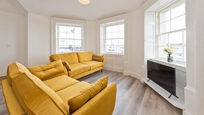 Una sala de estar con dos sofás color mostaza, un televisor de pantalla plana y ventanas amplias con vistas a la ciudad.