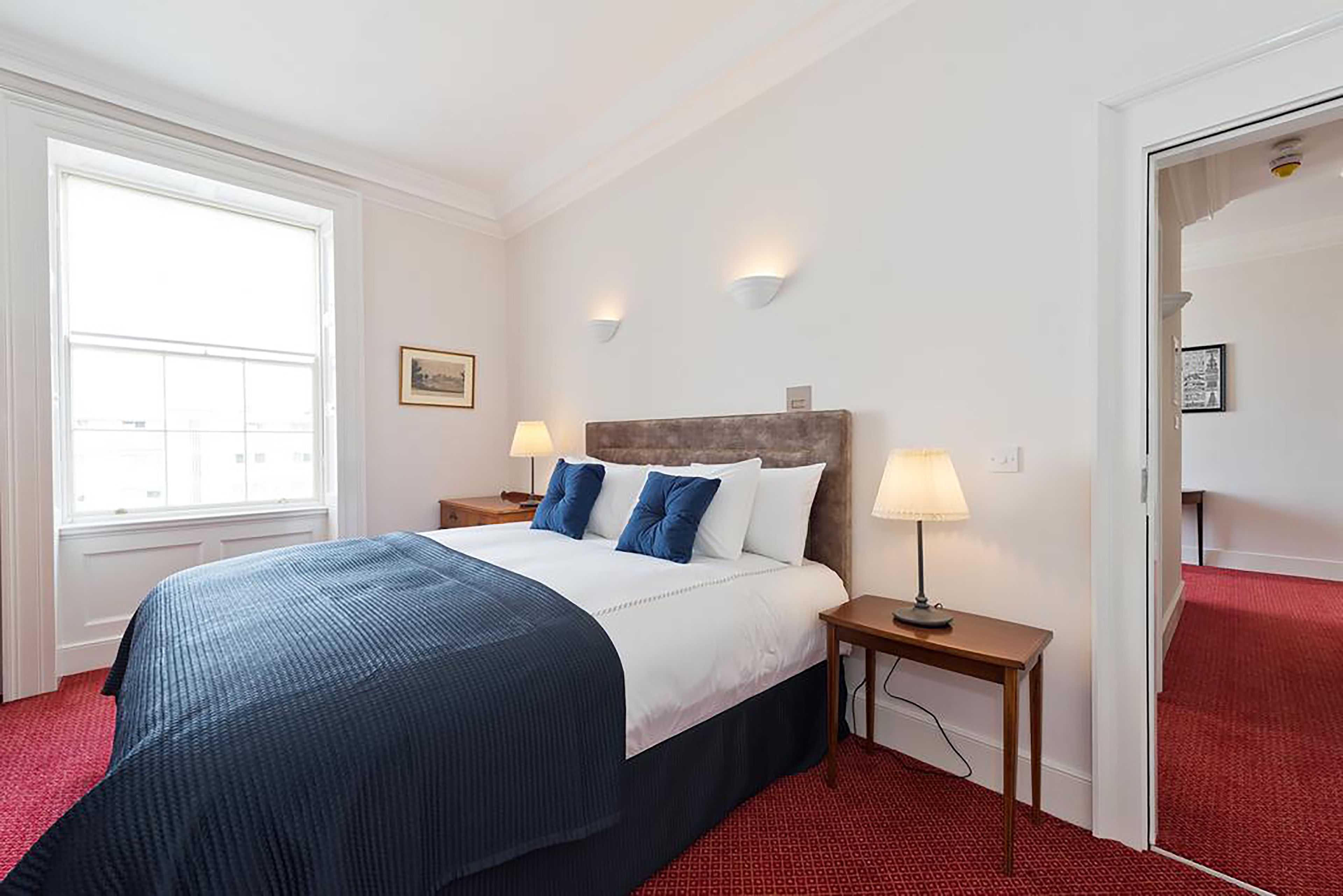 Quarto de hotel com uma cama grande, duas mesas de cabeceira com luminárias, uma janela com persianas e um tapete vermelho.