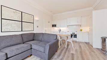 A modern living room with a gray sectional sofa, a dining area with a white table and chairs, and a kitchen with white cabinets and appliances.