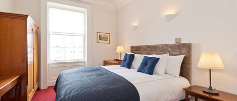 A hotel room with a large bed, two bedside tables with lamps, a wooden wardrobe, a window with blinds, and a framed picture on the wall.