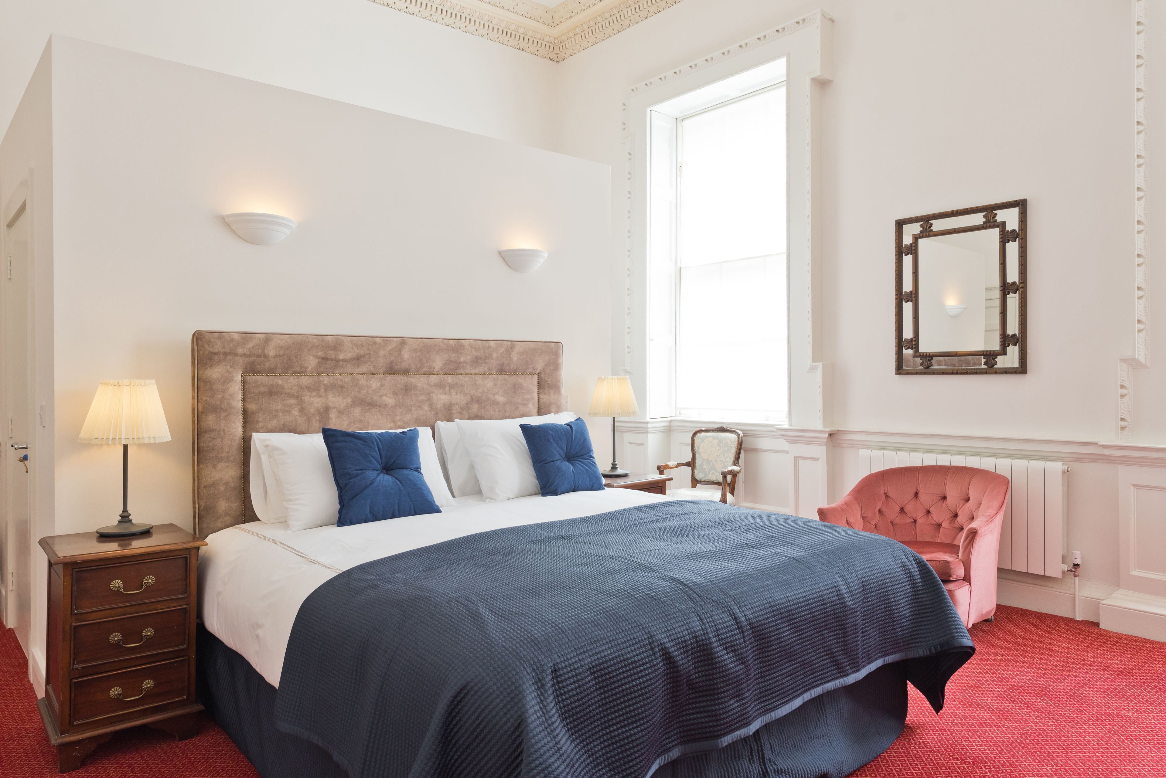 A bedroom with a large bed, two bedside tables with lamps, a pink armchair, and a large window.