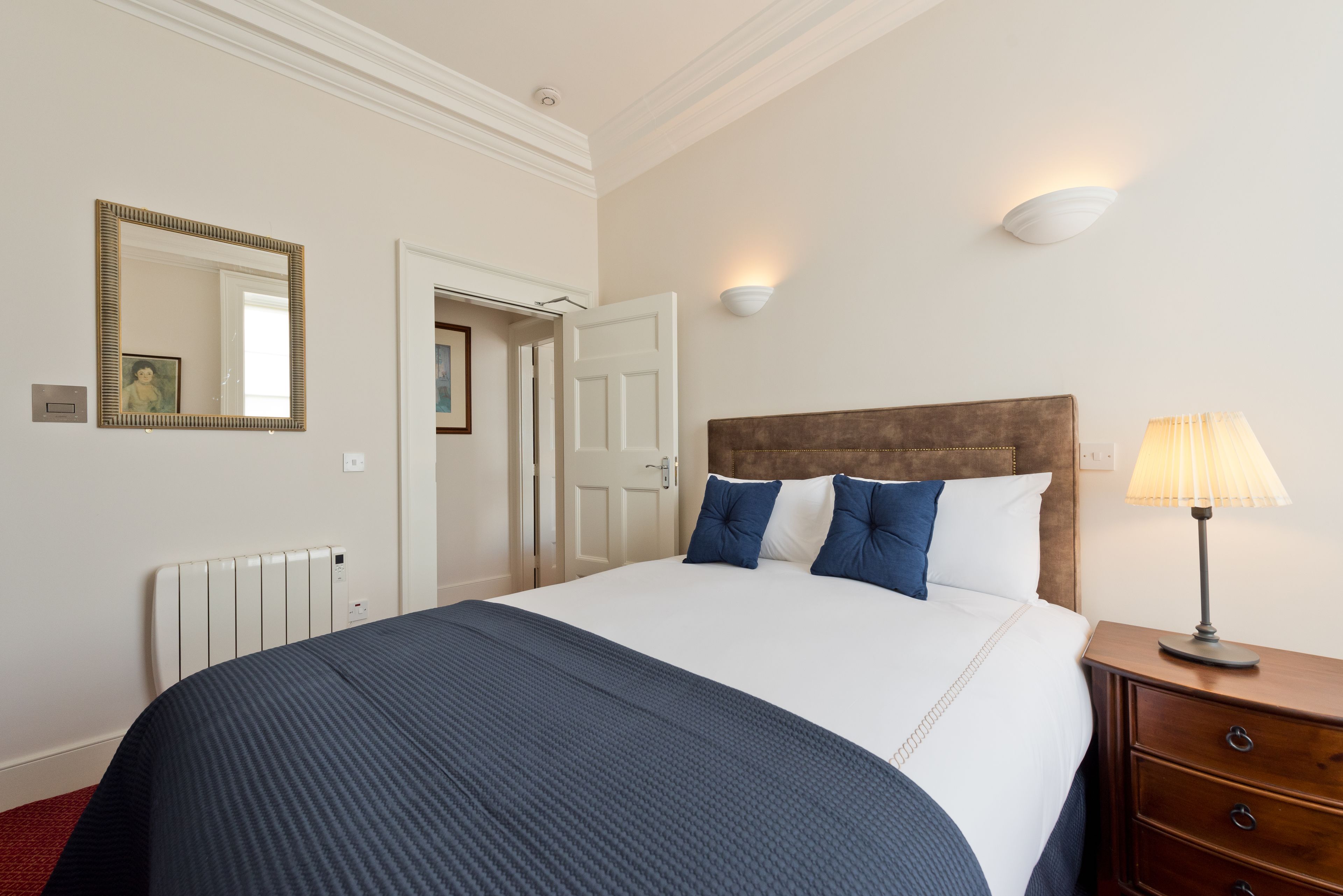 Habitación de hotel con una cama grande, una mesita de noche con lámpara, un espejo fijado en la pared y una puerta que da a otra habitación.
