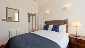 Habitación de hotel con una cama grande, una mesita de noche con lámpara, un espejo fijado en la pared y una puerta que da a otra habitación.