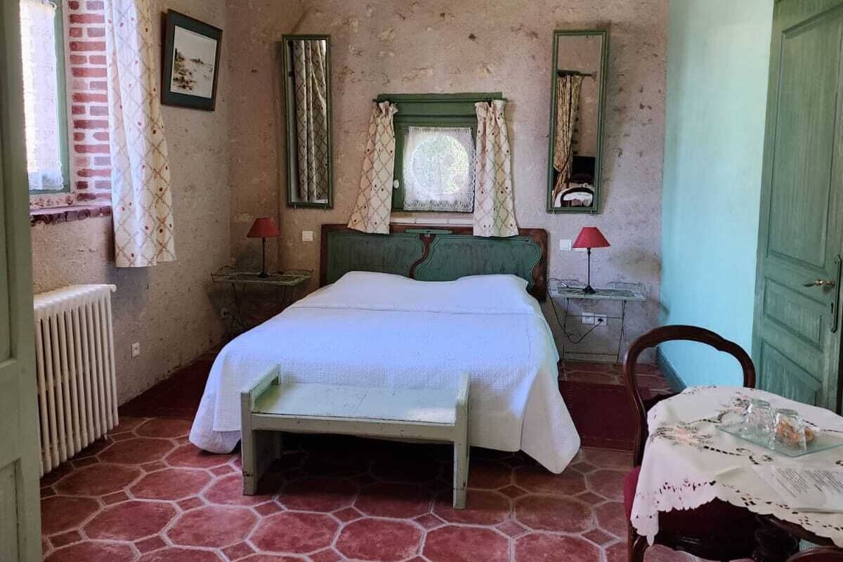 Une chambre à coucher comprenant un lit, une table, une chaise, une fenêtre avec des rideaux, un radiateur et un lustre.