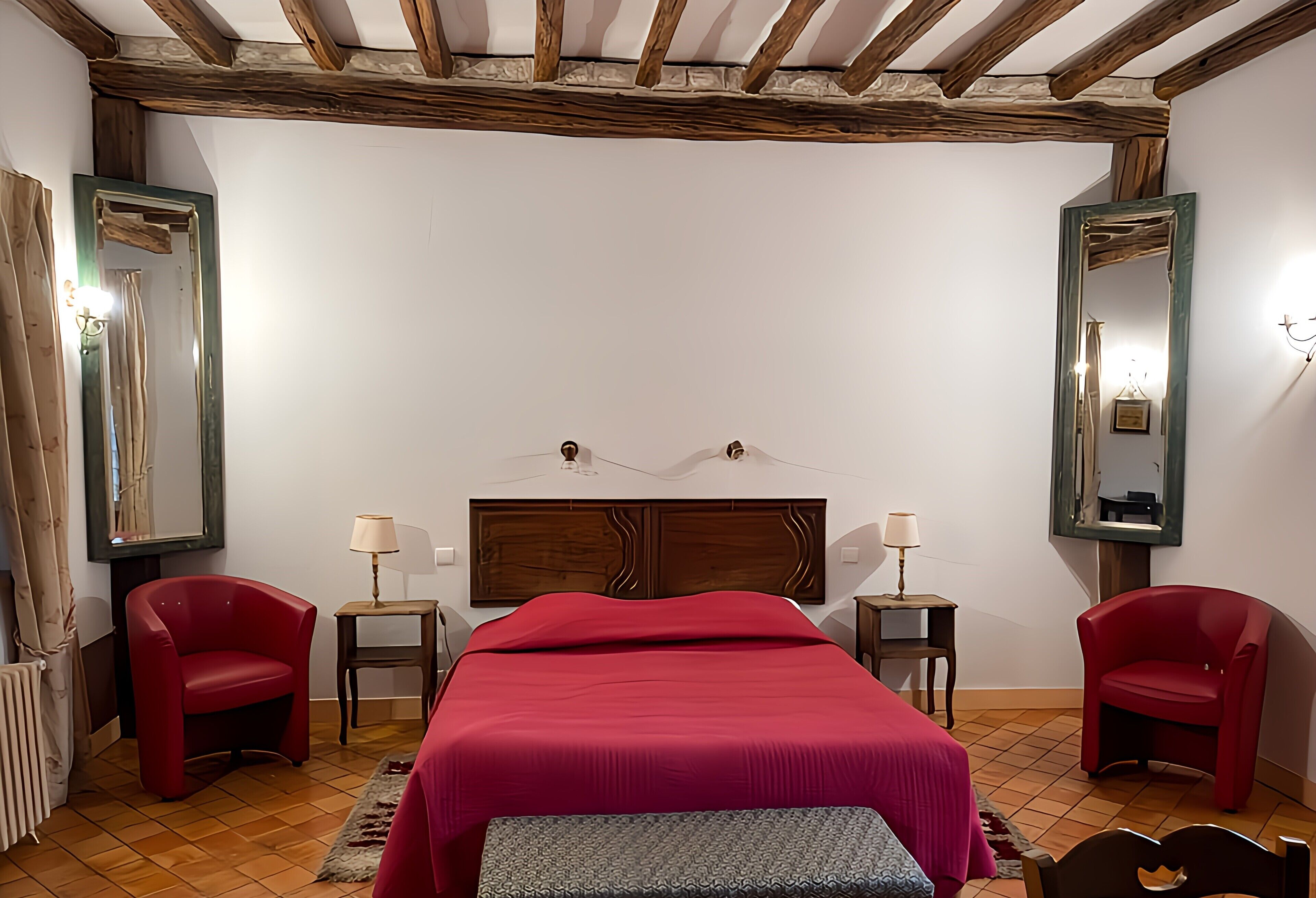 Un dormitorio con techo de madera, una cama grande con colcha roja, dos sillas rojas, dos mesitas de noche con lámparas y un espejo grande en la pared.
