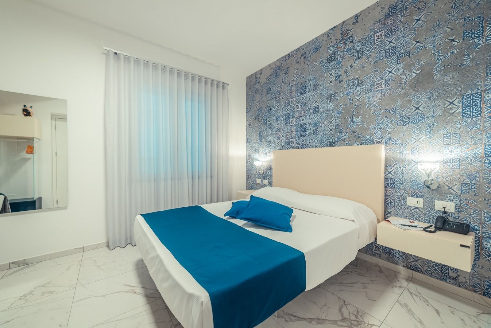 A hotel room with a bed, a nightstand, a mirror, and a patterned wall.