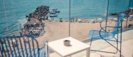 Une vue sur la plage depuis un balcon, avec une table et des chaises, donnant sur une plage de sable où l’on aperçoit des parasols et des gens.