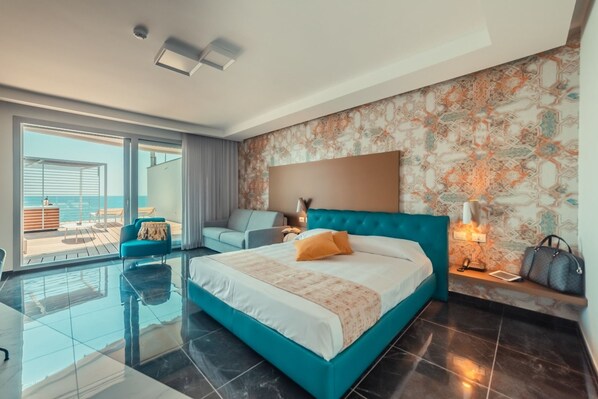 A modern hotel room with a large bed, a sofa, a balcony with a view of the sea, and a patterned wall.