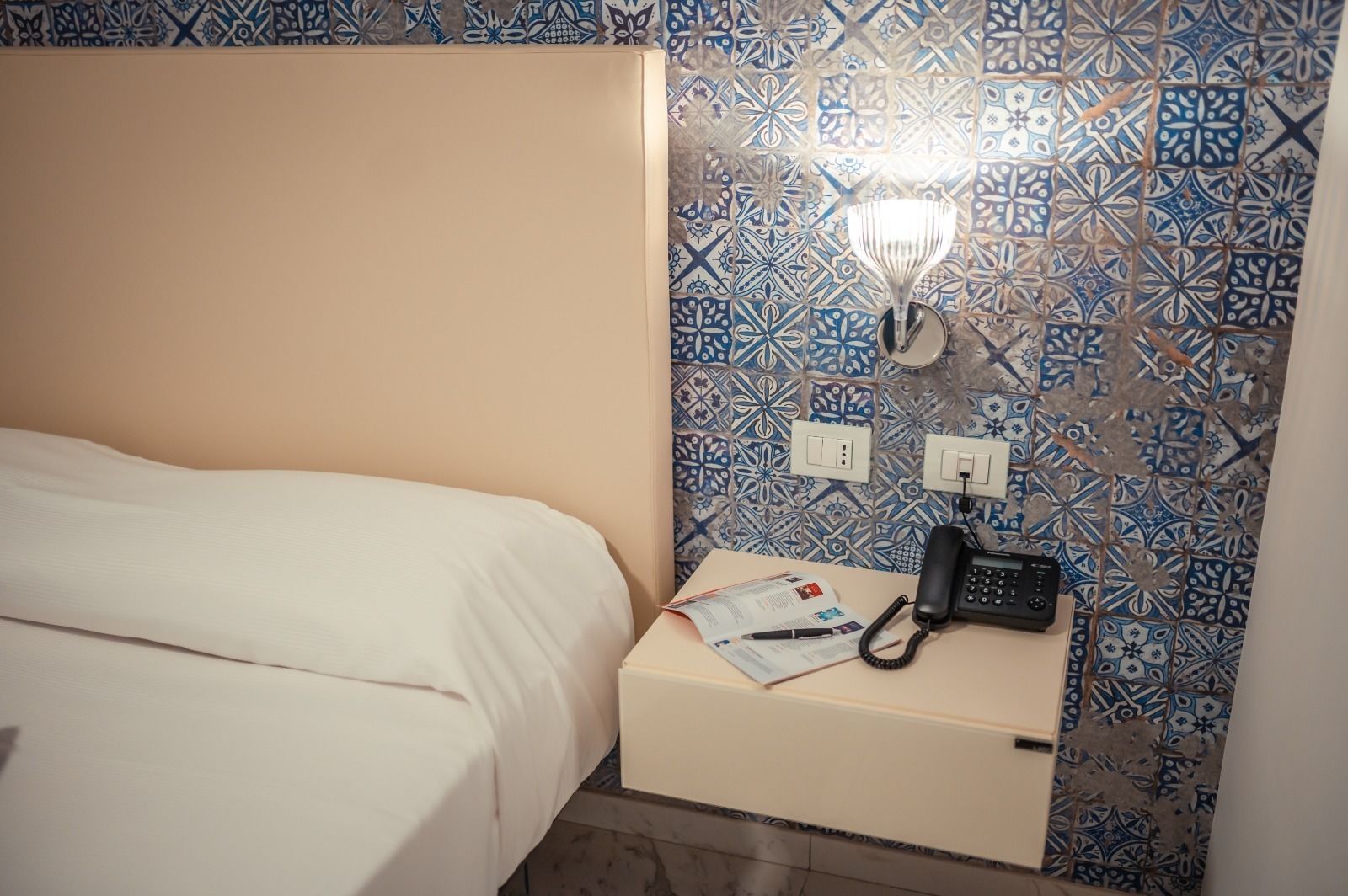 A hotel room with a bed, a nightstand with a lamp, a telephone, and a wall with patterned tiles.