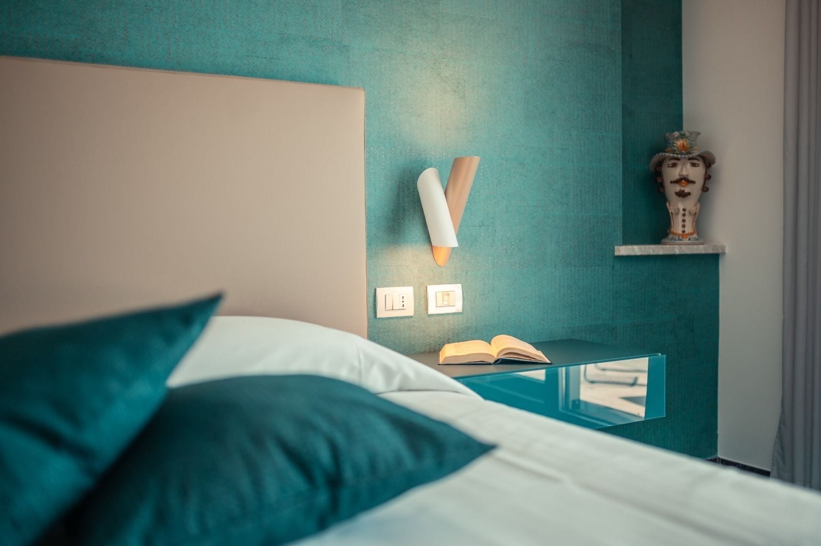 Een net opgemaakt bed met teal kussens, een nachtkastje met een open boek, en een wandlamp.