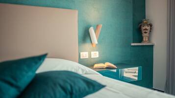 Een net opgemaakt bed met teal kussens, een nachtkastje met een open boek, en een wandlamp.