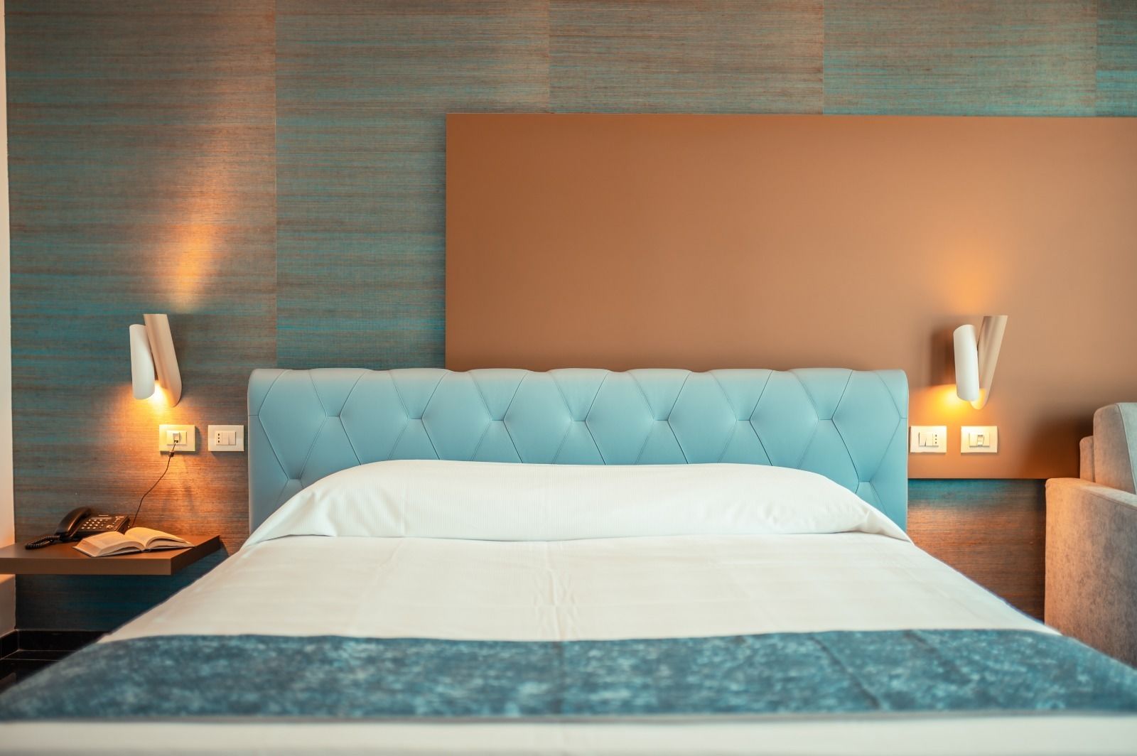 Quarto de hotel moderno com cabeceira estofada, mesa de cabeceira com abajur e criado-mudo com telefone e livro.
