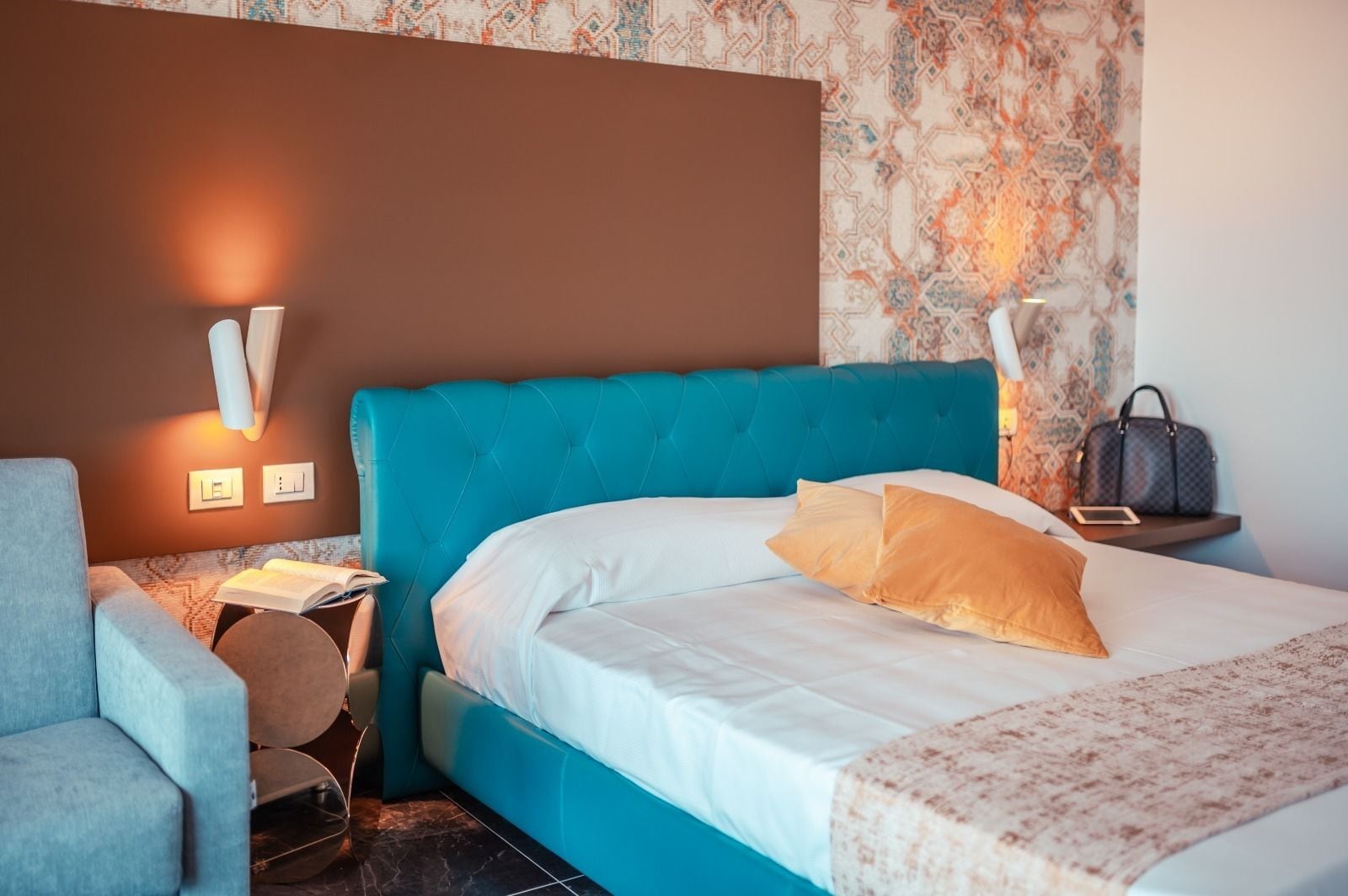 Habitación de hotel con una cama tapizada en color verde azulado, ropa de cama blanca, pared con estampado, una mesita de noche con lámpara y un sillón gris.