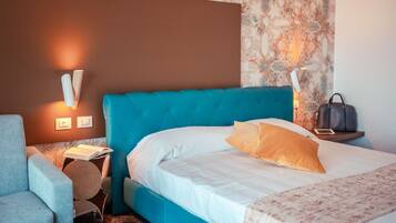 A hotel room with a teal upholstered bed, white bedding, a patterned wall, a side table with a lamp, and a grey armchair.