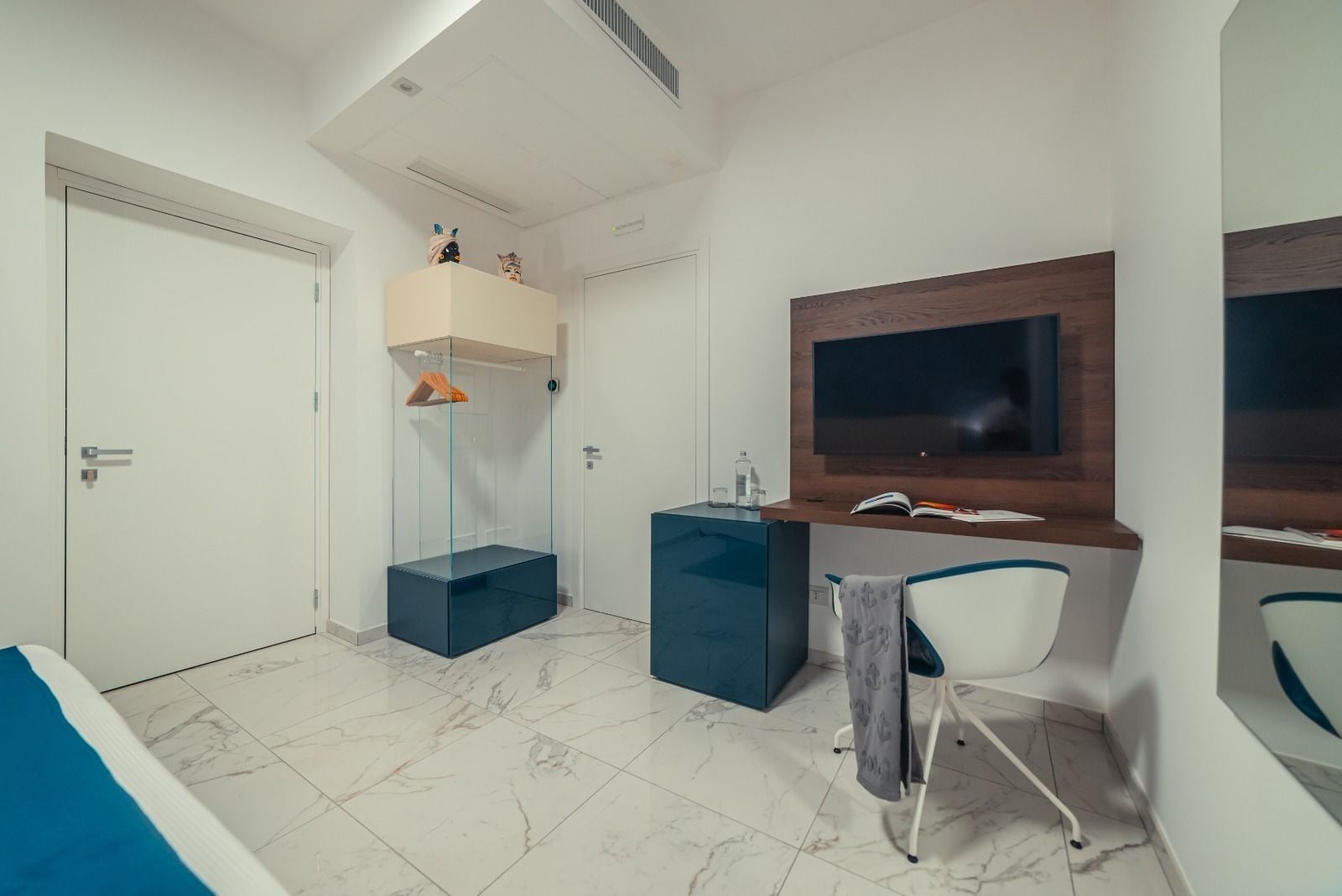 A modern bedroom with a bed, a desk, a chair, a TV, and a glass-enclosed wardrobe.
