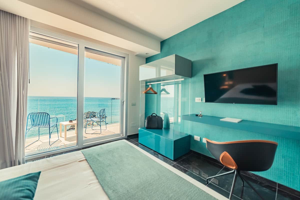 A modern hotel room with a large sliding glass door leading to a balcony with a view of the sea. There is a bed with a green throw, a desk with a chair, and a flat-screen TV mounted on the wall.