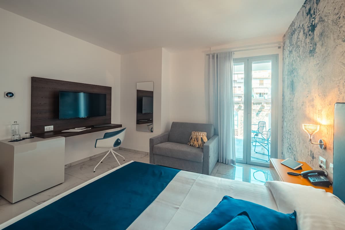 A modern hotel room with a bed, a desk with a TV, a chair, and a balcony with a view.