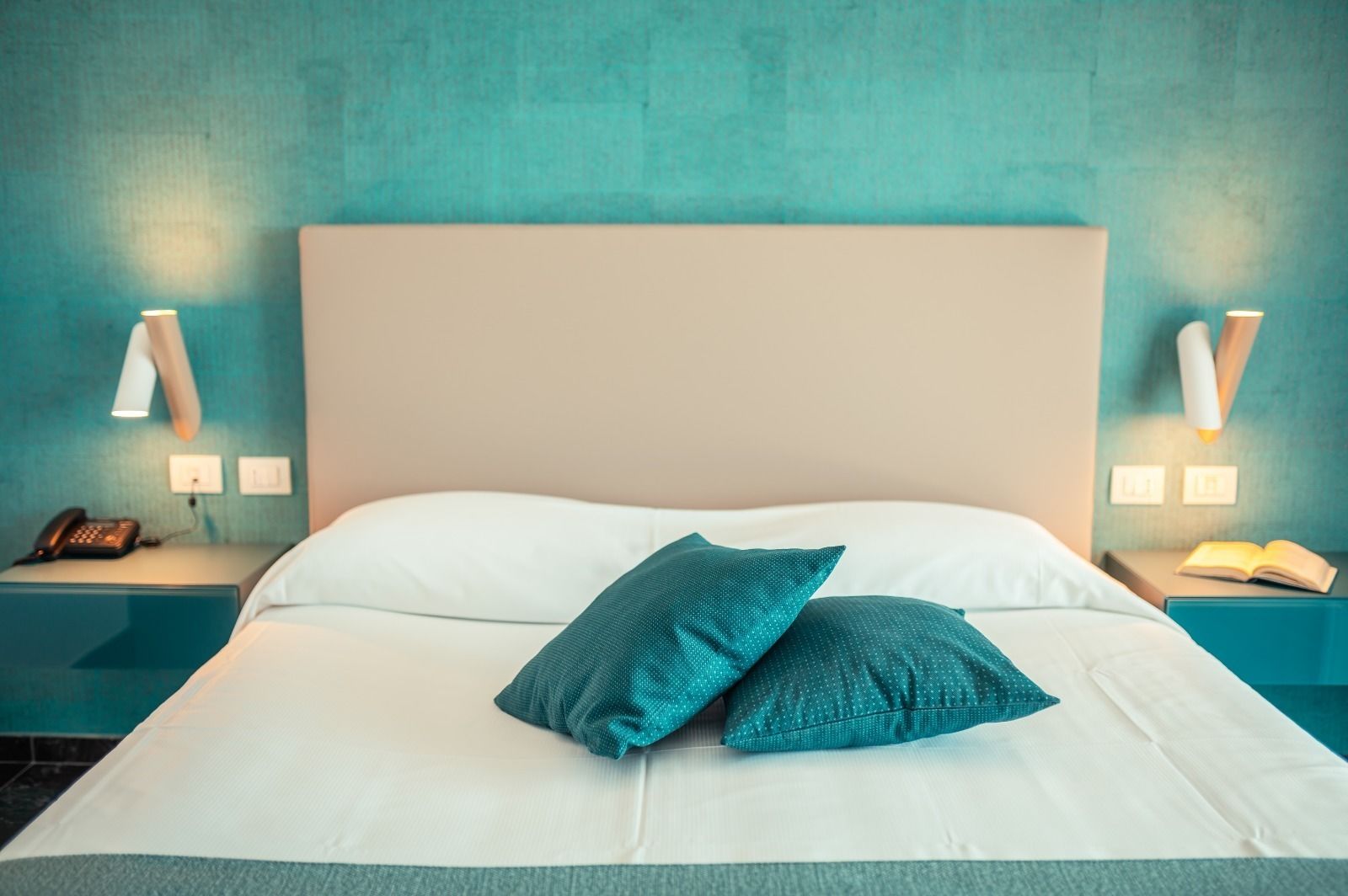 Una cama con ropa blanca, almohadillas turquesa, cabecera color beige y mesitas de noche turquesa con lámparas y un teléfono.