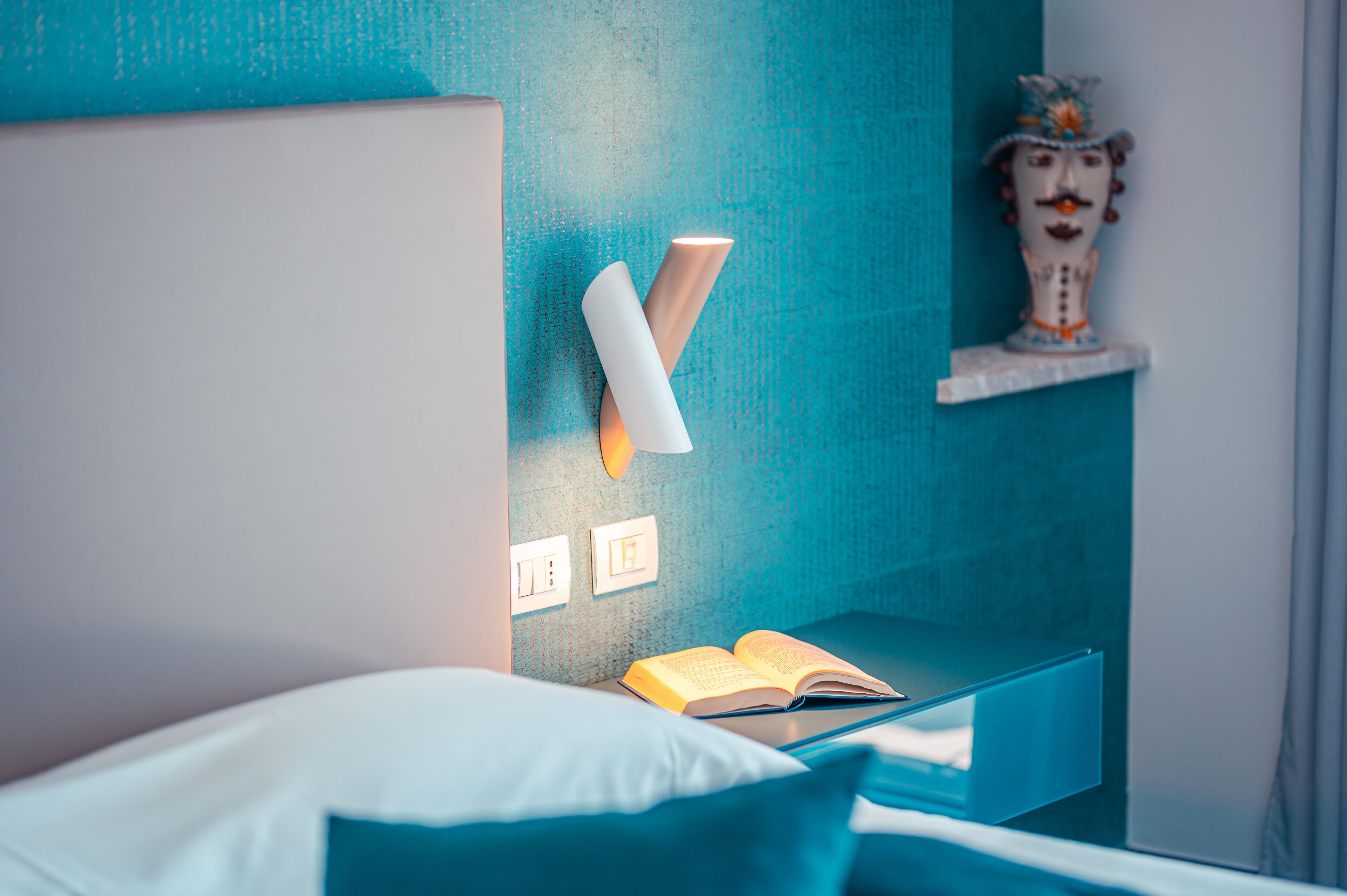 Een moderne slaapkamer met een blauwgroene accentmuur, een bedlampje, een decoratieve vaas en een open boek op het nachtkastje.