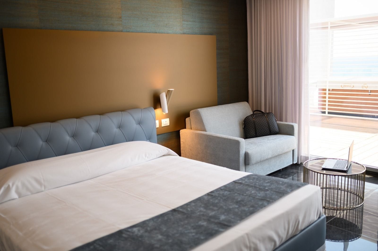 Habitación de hotel con una cama grande, un sillón gris, una mesita redonda con un ordenador portátil y vistas al exterior a través de amplias ventanas.