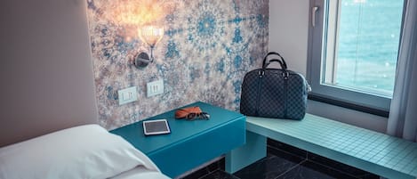 Una habitación de hotel con una cama, una mesita de noche con una tableta y gafas de sol, un bolso a cuadros, y vistas al mar a través de una ventana.