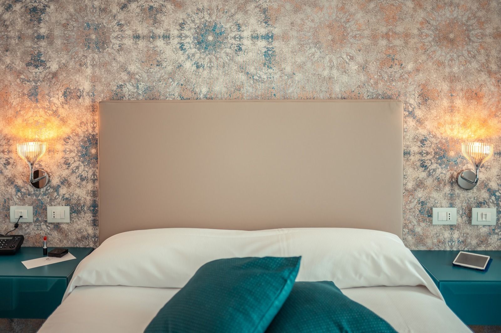 A bed with a beige headboard, teal nightstands, and a textured wall.