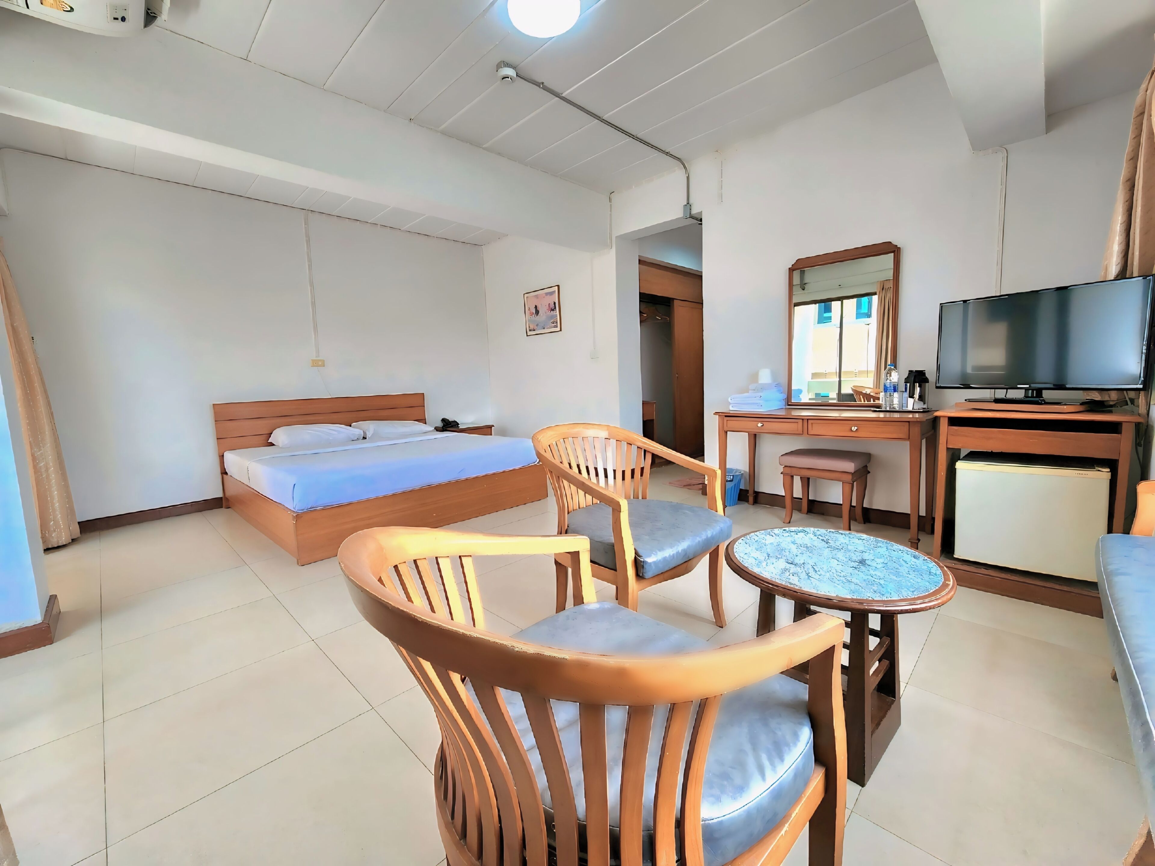A hotel room with a bed, wooden chairs, a small table, a desk with a television, and a refrigerator.