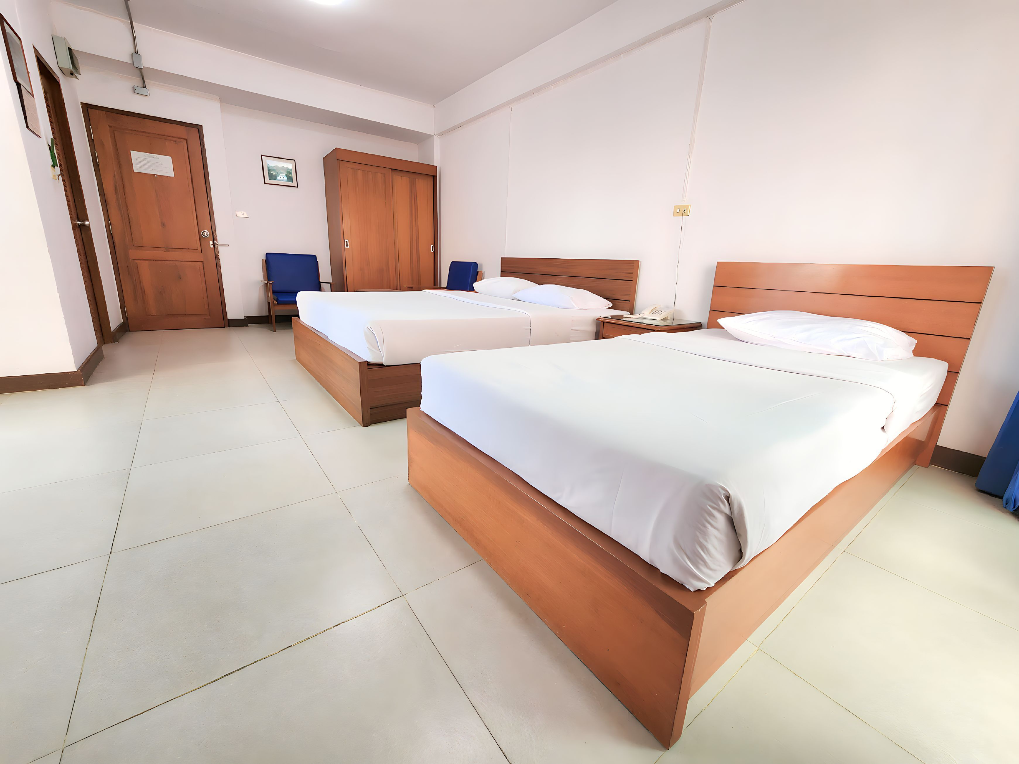 A hotel room with two wooden beds, a wooden wardrobe, a blue chair, and a bedside table with a phone.