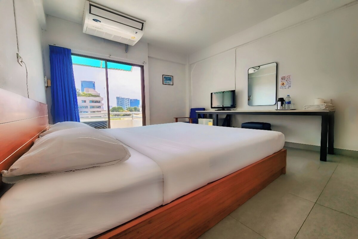 A hotel room with a bed, a desk with a computer, a TV, and a window with a view of buildings.