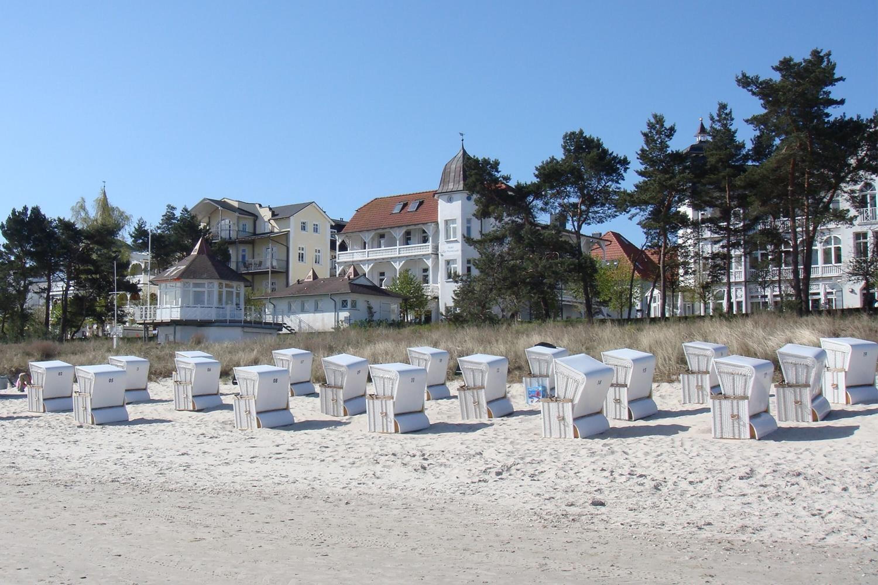 Un hotel frente a la playa con varias edificaciones, incluyendo una estructura blanca destacada con techo rojo. Filas de sillas de playa están dispuestas en la arena frente al hotel.